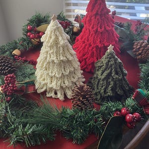 May include: Three handmade crochet Christmas trees in cream, red, and green, each topped with a wooden star. The trees are arranged on a red tablecloth surrounded by pine garland, pinecones, and red berries, creating a festive holiday display.