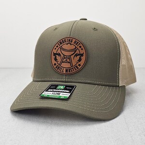 May include: A green and tan trucker hat with a brown leather patch that says "Smoking Hot Grill Master" with a grill graphic.