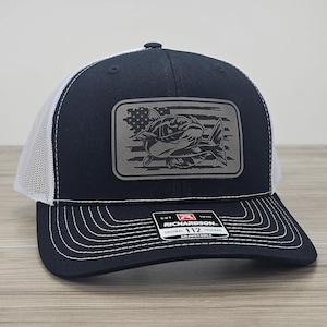 May include: A black and white trucker hat with a gray patch featuring a fish and an American flag design. The hat is adjustable and has the text "Richardson 112" on the back.