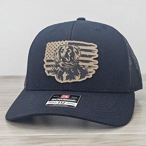 May include: Black trucker hat with a brown leather patch featuring a dog silhouette in front of an American flag design. The hat has a mesh back and the text "Richardson 112 Original Trucker Adjustable" on the label.