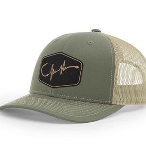 Puede incluir: Una gorra de camionero verde y beige con un parche negro que presenta un anzuelo dorado y un gr&aacute;fico de latido del coraz&oacute;n. El parche dice "Catch"