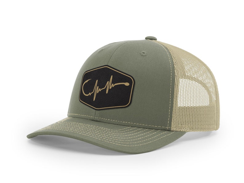 Puede incluir: Una gorra de camionero verde y beige con un parche negro que presenta un anzuelo dorado y un gr&aacute;fico de latido del coraz&oacute;n. El parche dice "Catch"