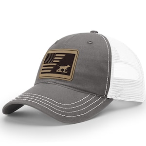 May include: A gray and white trucker hat with a brown patch featuring an American flag and a silhouette of a dog. The patch is sewn onto the front of the hat.