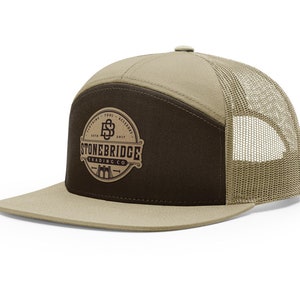 May include: A tan and brown trucker hat with a flat brim. The front panel is brown with a circular logo that reads "STONEBRIDGE TRADING CO." The hat has a mesh back and a snap closure.