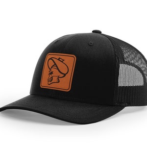 May include: Black trucker hat with a brown leather patch featuring a skull wearing a hat.