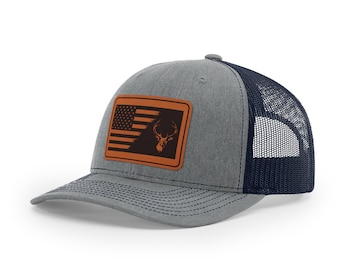 May include: A gray and navy blue trucker hat with a brown leather patch featuring an American flag with a deer silhouette.