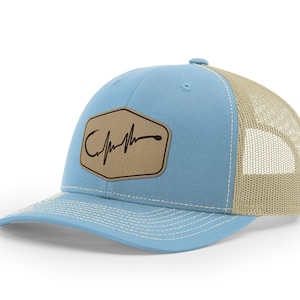 Puede incluir: Una gorra de camionero azul claro y beige con un parche de cuero marr&oacute;n que presenta un anzuelo de pesca y un gr&aacute;fico de latido del coraz&oacute;n. El parche tiene el texto "Catch the Feeling".