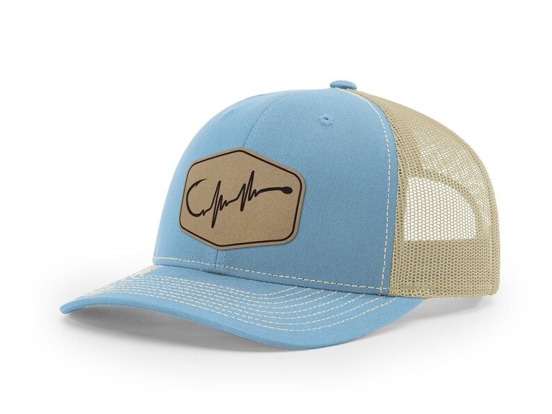 Puede incluir: Una gorra de camionero azul claro y beige con un parche de cuero marr&oacute;n que presenta un anzuelo de pesca y un gr&aacute;fico de latido del coraz&oacute;n. El parche tiene el texto "Catch the Feeling".