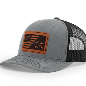 May include: A gray and black trucker hat with a brown leather patch featuring a stylized American flag with flying ducks.