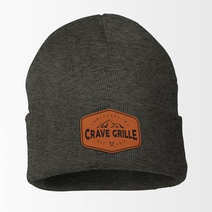 May include: A dark gray beanie with a brown leather patch that says "Vancouver, WA Crave Grille Est. 2016" with a mountain graphic and crossed utensils.