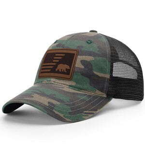 May include: A camouflage trucker hat with a brown leather patch featuring an American flag and a bear silhouette.