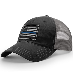 May include: A black and gray trucker hat with a distressed American flag patch featuring a thin blue line. The hat has a mesh back.