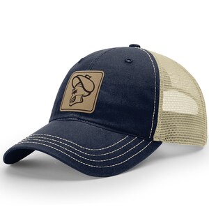 May include: Navy blue trucker hat with a tan mesh back and a curved brim. The hat features a square, tan leather patch with a skull wearing a turban. White stitching accents the brim.