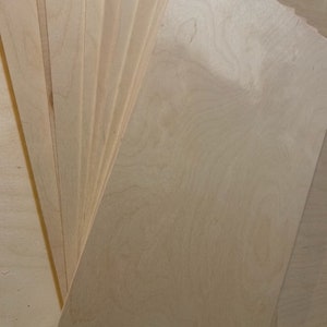 May include: A stack of thin, light wood sheets, likely plywood, with a natural wood grain pattern. The sheets are arranged in a fan shape, with the top sheet extending out to the right.