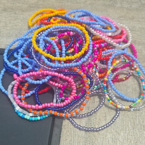 Puede incluir: Un montón grande de pulseras de cuentas de colores en tonos de azul, rosa, amarillo, naranja y morado. Las pulseras están hechas de cuentas pequeñas y redondas y están dispuestas en un patrón aleatorio.