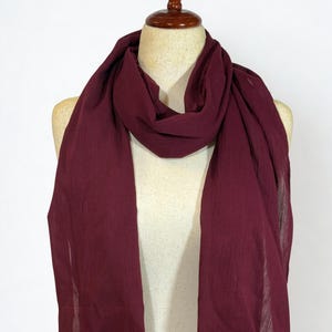 May include: A burgundy scarf draped over a mannequin. The scarf is made of a lightweight fabric with a slightly textured appearance. The ends of the scarf have a fringed edge. The scarf is a solid color and appears to be new.
