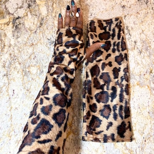 以下が含まれることがあります： ヒョウ柄の柄が入ったアームウォーマーのペア。アームウォーマーはタン色で、濃い茶色の斑点があり、柔らかく毛皮のような素材で作られています。明るい茶色の表面に表示されています。