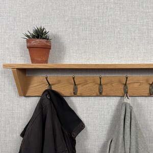 Op de afbeelding: Een lichtbruine houten wandplank met vijf antieke messingkleurige metalen haken bevestigd aan een horizontale achterplaat. Een kleine groene vetplant in een terracotta pot staat op de plank, terwijl een zwarte jas en een grijze trui eronder hangen.