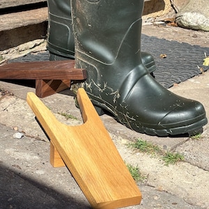 May include: Two wooden boot jacks, one crafted from light-colored oak and the other from dark walnut, positioned on a concrete surface. Each boot jack features a U-shaped cutout at the top end for removing footwear, with a small support block underneath.