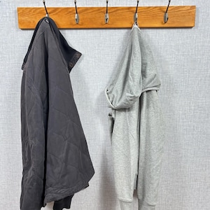 May include: A dark charcoal gray quilted jacket with a high collar and a light gray hooded sweatshirt hanging on a wooden rack with five metal hooks. The garments are suspended against a light gray textured wall surface.