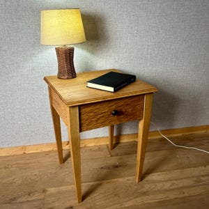 May include: A wooden bedside table with a single drawer and a small lamp. The table is made of light-coloured wood and has a dark knob on the drawer. A book with a black cover rests on top. The lamp has a woven base and a beige lampshade.