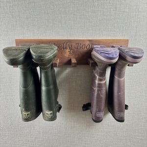 Könnte beinhalten: Ein hölzerner Stiefelhalter mit dem eingravierten Text "Welly Boots" hält zwei Paar Gummistiefel. Ein Paar ist dunkelgrün, das andere ist ein gesprenkeltes Lila. Der Ständer ist aus dunklem Holz und an einer hellgrauen Wand befestigt.