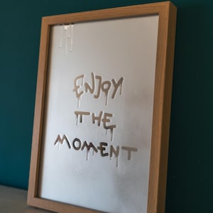 Op de afbeelding: Een ingelijst kunstwerk met de woorden "ENJOY THE MOMENT" geëtst in een wit oppervlak. De tekst heeft een druipend effect en de lijst is van lichtgekleurd hout. De achtergrond is donkerblauwgroen.