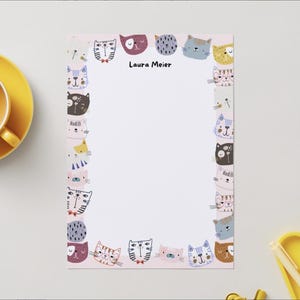 Könnte beinhalten: Hellrosa Notizblock mit einer Bordüre aus bunten Cartoon-Katzenköpfen. Der Name "Laura Meier" ist oben aufgedruckt. Eine gelbe Tasse und Untertasse befinden sich in der oberen linken Ecke.