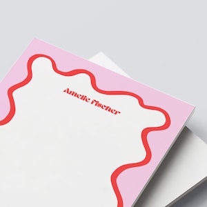 Könnte beinhalten: Ein Stapel Notizblöcke mit einem rosa Rand und einem roten Wellendesign. Der Text "Amelie Fischer" ist rot auf dem weißen Notizblock gedruckt. Der Hintergrund ist hellgrau.