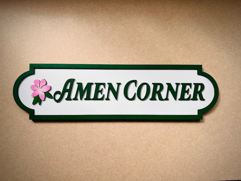 May include: A white sign with green trim and a pink flower design. The sign reads "AMEN CORNER".
