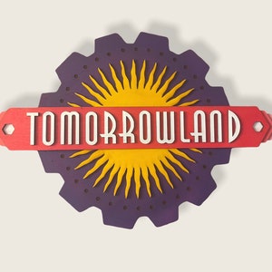 May include: A purple and yellow sign with the text "TOMORROWLAND" in white letters. The sign is shaped like a gear with a sunburst design in the center.