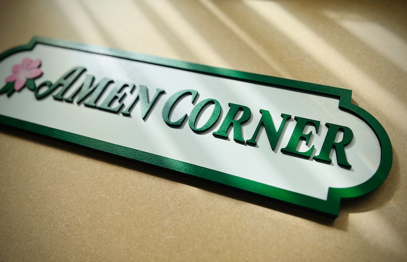 May include: A green and white wooden sign with the text "AMEN CORNER" in green raised letters. The sign has a decorative border and a small pink flower design.