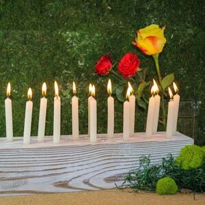 May include: A white wooden candle holder with 12 lit white candles. The holder is sitting on a brown surface with green moss and a yellow and red rose.