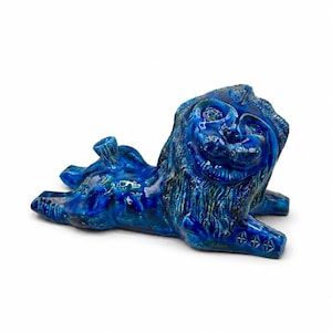 Op de afbeelding: Een blauw geglazuurde keramische leeuwenfiguur in liggende houding. De sculptuur heeft gedetailleerde gelaatstrekken en een getextureerde manen. De poten van de leeuw zijn versierd met stervormige patronen.