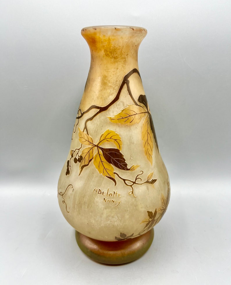 May include: A pale yellow glass vase with a pearlescent finish and a floral design in shades of brown and gold. The vase is signed "ADELLOTTE NANCY".