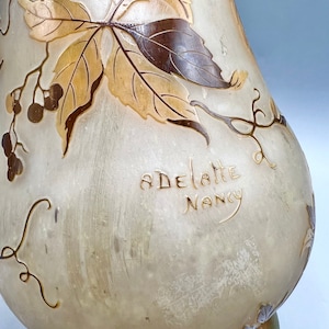 May include: A white glass vase with a pear-shaped body and a brown and gold floral design. The vase is signed "ADELLATE NANCY".