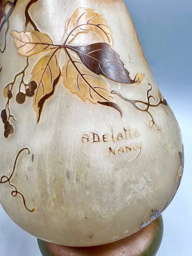 May include: A white glass vase with a pear-shaped body and a brown and gold floral design. The vase is signed "ADELLATE NANCY".