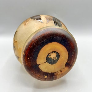 May include: A round, cream-colored ceramic vase with a brown wooden base. The vase has a carved design of leaves and flowers.