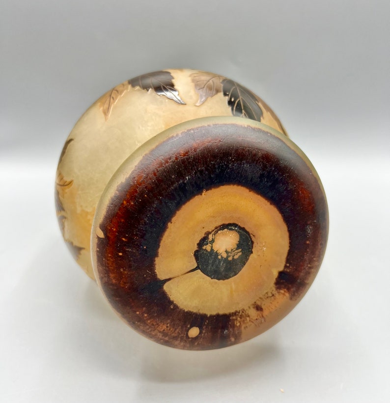 May include: A round, cream-colored ceramic vase with a brown wooden base. The vase has a carved design of leaves and flowers.