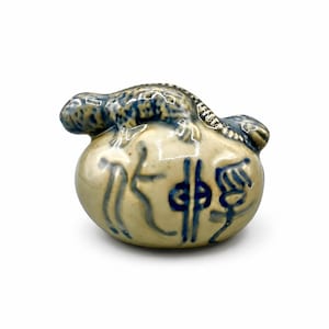 May include: A small, ceramic figurine of a pouch with a lizard on top. The pouch is cream-colored with blue painted details and symbols. The lizard is blue with darker blue spots. The figurine is likely a decorative item.