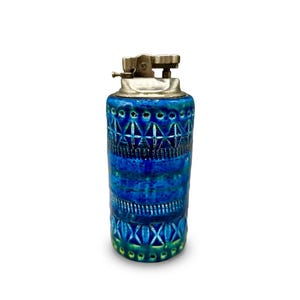 Op de afbeelding: Een vintage keramische aansteker met een cilindrische vorm. De aansteker heeft een levendig blauw glazuur met geometrische patronen en een zilverkleurige metalen bovenkant. De basis van de aansteker heeft een groene tint.