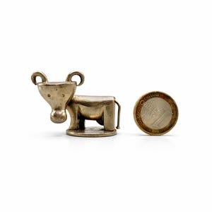 May include: A small, silver-colored metal cow figurine with a rounded head, curved horns, and a rectangular body. The cow stands on a circular base. A one Euro coin is next to the cow for size comparison.
