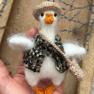 May include: A handmade white goose figurine with an orange beak and feet, wearing a brown tweed vest and hat. The goose has a beige shoulder bag. The figurine is a charming, whimsical piece.