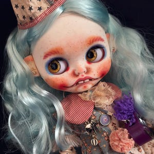 Op de afbeelding: Een op maat gemaakte kunstdoll met blauw haar en een grillig clownachtig uiterlijk. De pop heeft een geschilderd gezicht met sproeten, grote ogen en een kleine hoed versierd met sterren. De outfit van de pop omvat een jurk met bloemenprint en diverse decoratieve elementen.