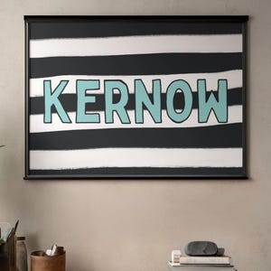 May include: Framed artwork featuring the word "KERNOW" in light blue, set against a backdrop of black and white horizontal stripes. The artwork is displayed in a black frame, creating a bold contrast. The overall aesthetic is modern and graphic.