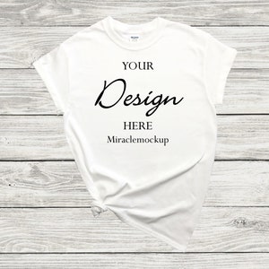 K&ouml;nnte beinhalten: Wei&szlig;es T-Shirt-Mockup mit dem Text "YOUR Design HERE Miraclemockup" auf wei&szlig;em Holz Hintergrund.