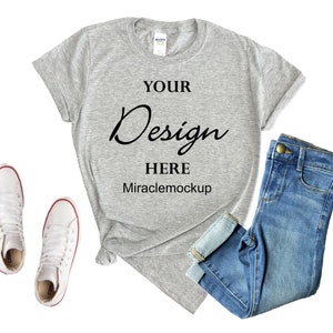 May include: A heather grey t-shirt with the text "YOUR Design HERE Miraclemockup" on the front. The t-shirt is laid flat on a white background with a pair of white high-top sneakers and a pair of blue jeans.