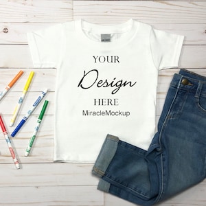 May include: White t-shirt with the text "YOUR Design HERE MiracleMockup" on a white wooden background with a pair of blue jeans and colorful markers.
