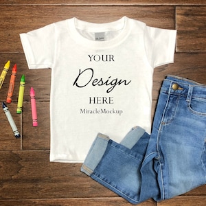 Könnte beinhalten: Ein weißes T-Shirt und eine blaue Jeans liegen auf einer Holzoberfläche, zusammen mit bunten Wachsmalstiften. Auf dem T-Shirt steht der Text "YOUR Design HERE MiracleMockup". Die Jeans ist gefaltet, und die Stifte sind gelb, rot, grün, rosa und blau.