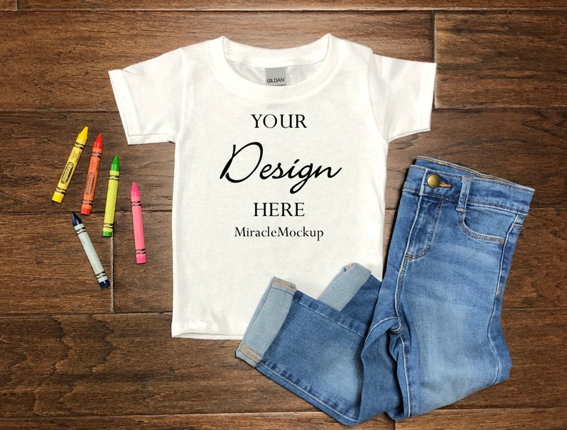 May include: A white t-shirt and blue jeans are displayed on a wooden surface, alongside colorful crayons. The t-shirt has the text "YOUR Design HERE MiracleMockup". The jeans are folded, and the crayons are in yellow, red, green, pink, and blue.